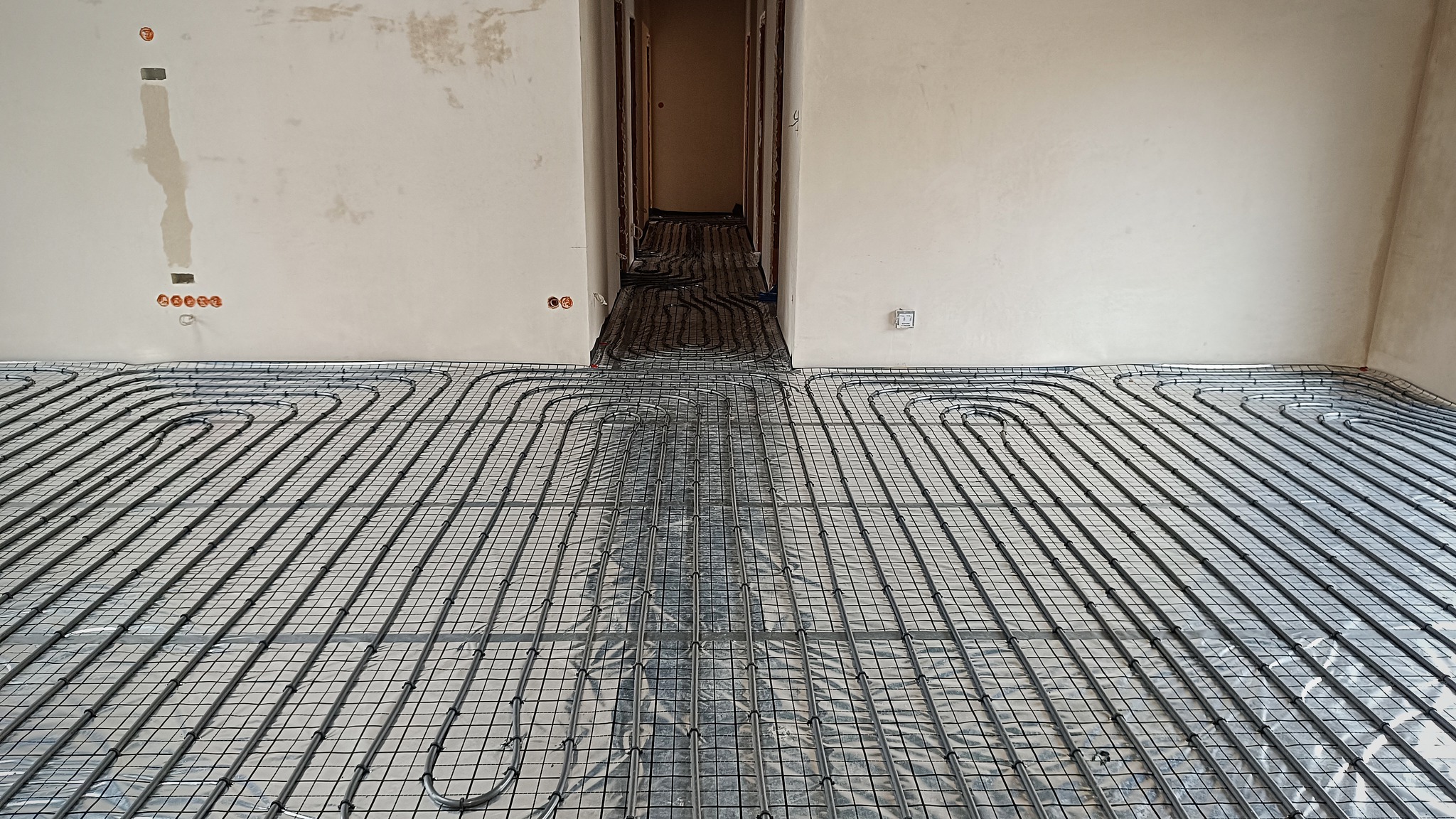 Rozłożona instalacja ogrzewania podłogowego na folii izolacyjnej w surowym pomieszczeniu z widocznymi puszkami elektrycznymi na ścianie i przejściem do kolejnego pokoju.
