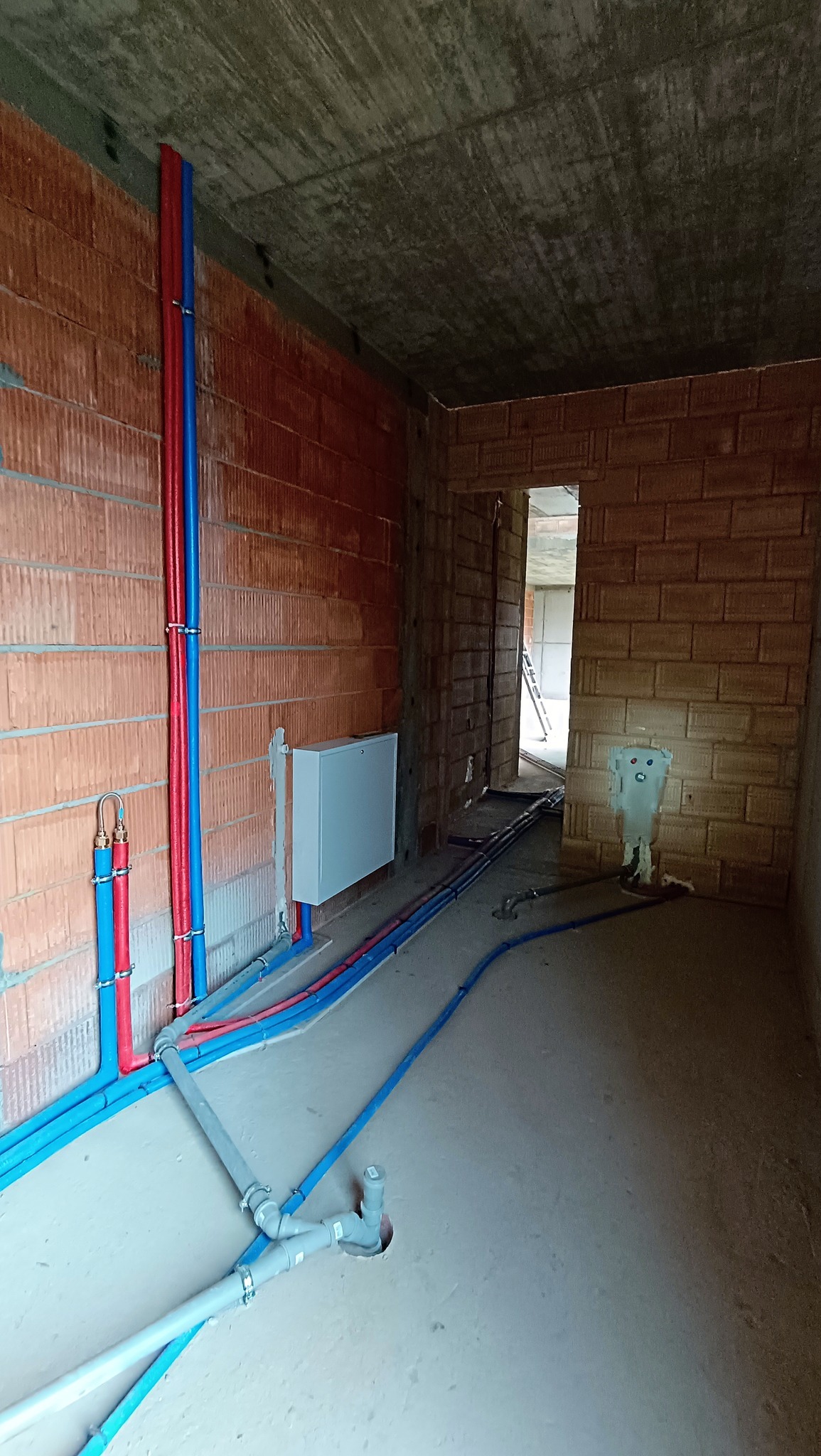 Wnętrze w trakcie instalacji wodno-kanalizacyjnej: widoczne rury w kolorach niebieskim i czerwonym prowadzone wzdłuż ściany z cegły, szara skrzynka na ścianie, fragment instalacji kanalizacyjnej...