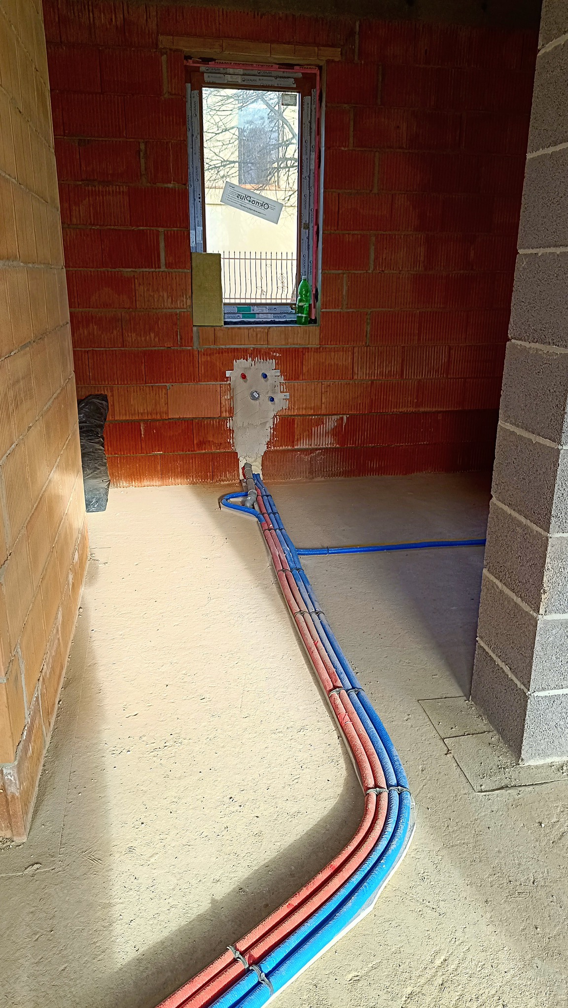 Wnętrze w trakcie budowy z widocznymi rurami instalacji wodnej i grzewczej, prowadzącymi od ściany z oknem do podłogi. Rury w kolorach niebieskim i czerwonym, zabezpieczone opaskami. Ściany...