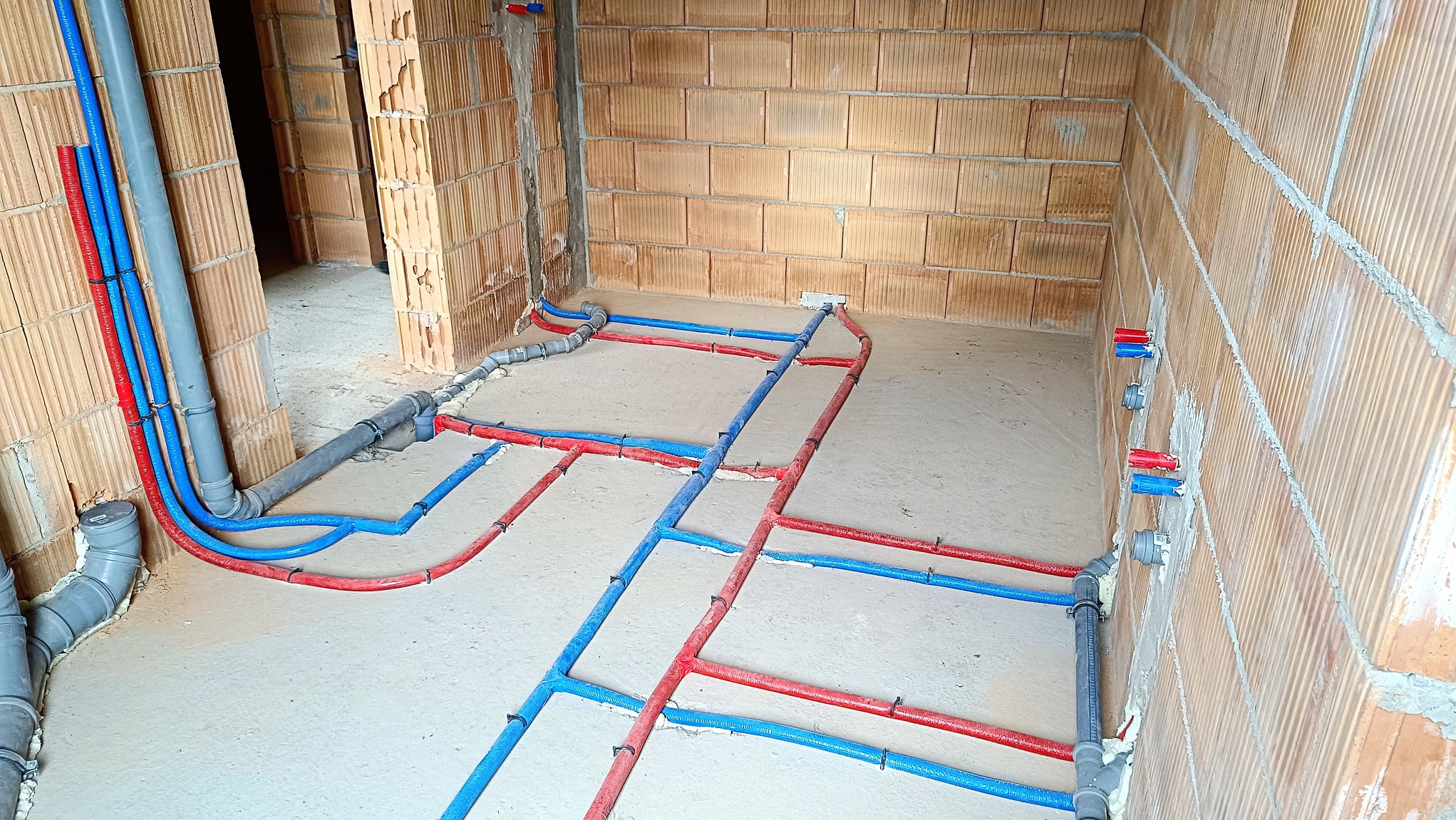 Wnętrze w trakcie budowy z widoczną instalacją wodno-kanalizacyjną: rury ciepłej i zimnej wody (czerwone i niebieskie) prowadzone po podłodze i ścianach z bloczków ceramicznych, oraz rury odpływowe...