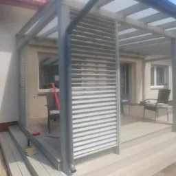 Nowoczesna, szara pergola z żaluzjami na tarasie przylegającym do domu jednorodzinnego, widok z zewnątrz na częściowo umeblowaną przestrzeń wypoczynkową.
