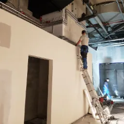 Pracownik na drabinie wykańcza ścianę w pomieszczeniu z widoczną instalacją pod sufitem, drugi pracownik w tle.