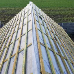 Konstrukcja dachu z drewnianych łat, pokryta membraną dachową z widocznym nadrukiem producenta, na tle zielonego trawnika i betonowego ogrodzenia.