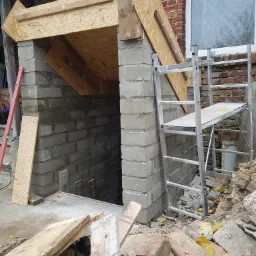 Murowana konstrukcja wejścia do piwnicy w trakcie budowy, widoczne szalunki z płyty OSB, rusztowanie i gruz.