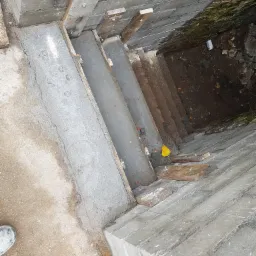 Wykop pod fundamenty z częściowo wylanym betonem, widoczne drewniane elementy szalunku mocowane śrubami, oraz fragment murowanej ściany z bloków betonowych.