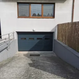 Szara segmentowa brama garażowa z trzema małymi okienkami, zamontowana w podziemnym wjeździe do garażu, widoczna z zewnątrz. Nad bramą okno z żaluzjami, po prawej stronie bambusowa osłona.