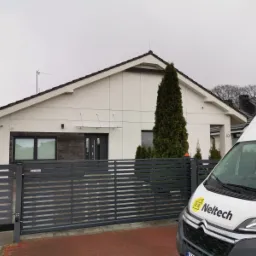 Nowoczesny dom jednorodzinny z szarą, poziomą bramą wjazdową, widoczny fragment firmowego samochodu dostawczego z logo po prawej stronie.