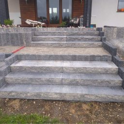 NaturaDOM - Schody wejściowe do domu wyłożone szarą kostką granitową, częściowo zasypane piaskiem, z widocznym poziomem na jednym ze stopni.