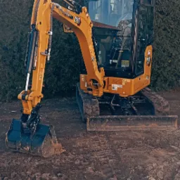 Żółta minikoparka CAT 302 CR z czarną kabiną i łyżką, stojąca na ziemi z widocznymi śladami gąsienic, na tle gęstego, zielonego żywopłotu.