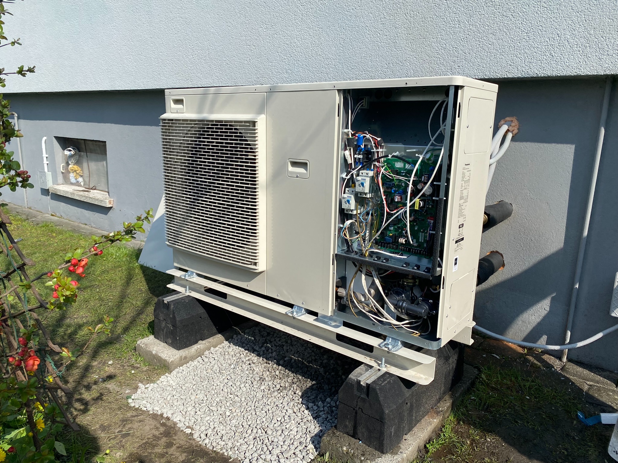 Zewnętrzna jednostka pompy ciepła Panasonic Aquarea z otwartym panelem bocznym, widoczne wewnętrzne podzespoły i instalacja na betonowych blokach, z rurami wchodzącymi do budynku.