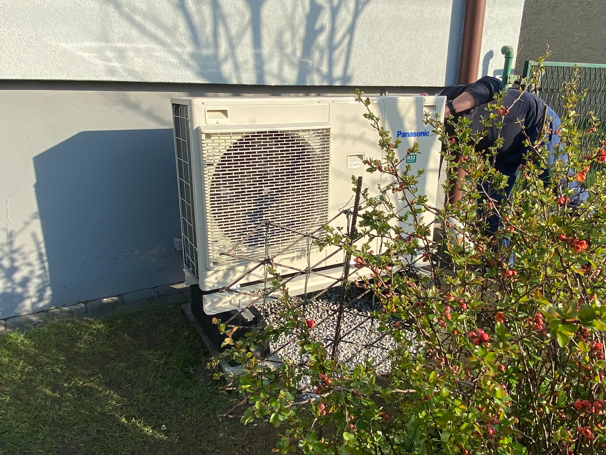 Zewnętrzna jednostka klimatyzacji Panasonic, zamontowana przy ścianie budynku, z widocznym pracownikiem instalującym urządzenie, częściowo zasłonięta przez krzew z czerwonymi pąkami.