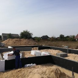 SAN-REMONT - Pracownik w niebieskim kombinezonie układa białe bloczki izolacyjne na czarnej hydroizolacji fundamentu domu jednorodzinnego w trakcie budowy, widoczne ułożone bloczki betonowe, piasek i domy w tle.