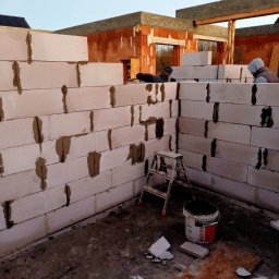 SAN-REMONT - Wznoszenie ścian z białych bloczków na budowie domu jednorodzinnego, widoczni dwaj pracownicy w odzieży roboczej, drabina, wiadro z zaprawą i porozrzucane fragmenty bloczków na betonowej posadzce.