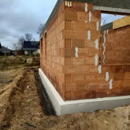 Wznoszone ściany z czerwonej ceramiki poryzowanej na fundamencie z betonu, widoczne spoiny pionowe wypełnione zaprawą, izolacja przeciwwilgociowa fundamentu, wykopany rów wzdłuż ściany, w tle domy...