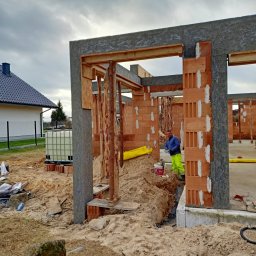 SAN-REMONT - Budowa domu jednorodzinnego: murowane ściany z pustaków ceramicznych z widocznymi otworami, szalunki z drewna i płyt OSB, pracownik w odblaskowej odzieży roboczej w tle, piasek i materiały...