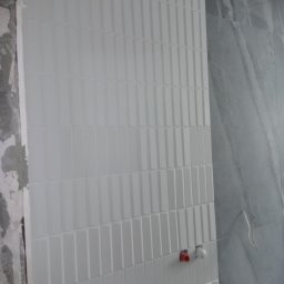 SAN-REMONT - Ściana w trakcie układania białych, prostokątnych płytek z delikatną fakturą; widoczne fragmenty surowej ściany z resztkami zaprawy oraz przyłącza wodne.