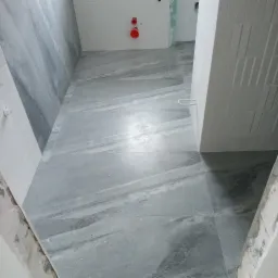 Wykończona podłoga w łazience z szarych płytek imitujących marmur, częściowo ułożona ściana z białych, wąskich płytek, widoczne fragmenty surowego muru przy futrynach drzwi.