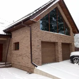 Elewacja budynku z brązowej cegły klinkierowej, z dużym trójkątnym oknem odbijającym drzewa, brązowe rynny, dwustanowiskowy garaż w zimowej scenerii ze śniegiem.