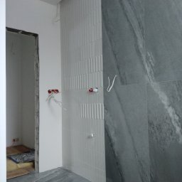 SAN-REMONT - Wnętrze łazienki w trakcie remontu: białe, pionowe płytki ułożone w pasy obok szarych płyt imitujących marmur, wystające kable elektryczne, surowa ościeżnica drzwi.