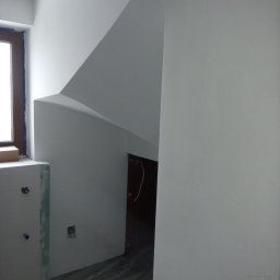 SAN-REMONT - Świeżo pomalowane na biało wnętrze z oknem, widoczne ślady po taśmie malarskiej, surowy stan wykończenia przy oknie i podłodze.