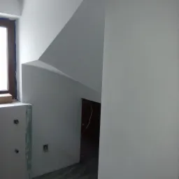 Świeżo pomalowane na biało wnętrze z oknem, widoczne ślady po taśmie malarskiej, surowy stan wykończenia przy oknie i podłodze.