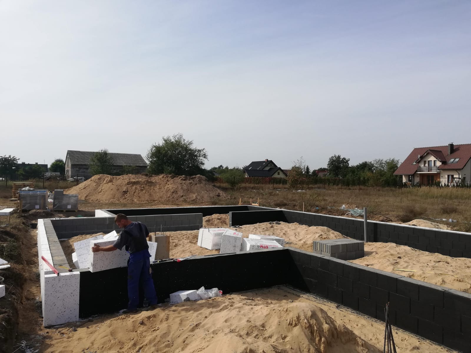 Pracownik w niebieskim kombinezonie układa białe bloczki izolacyjne na czarnej hydroizolacji fundamentu domu jednorodzinnego w trakcie budowy, widoczne ułożone bloczki betonowe, piasek i domy w tle.