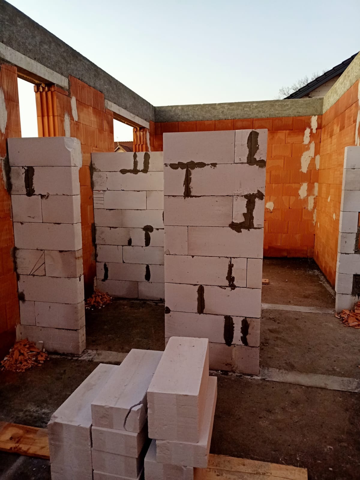 Wnętrze budynku w trakcie budowy: ściany z czerwonej cegły dziurawki i białe ściany działowe z betonu komórkowego, świeżo murowane, z widocznymi spoinami z zaprawy. Na podłodze stos białych bloczków.