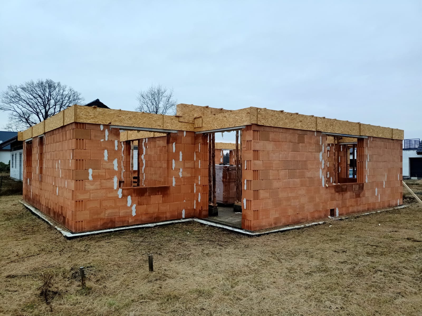 Surowy stan domu jednorodzinnego z czerwonej cegły, widoczne otwory okienne i drzwiowe, przygotowany pod wieniec, na tle jesiennego krajobrazu.