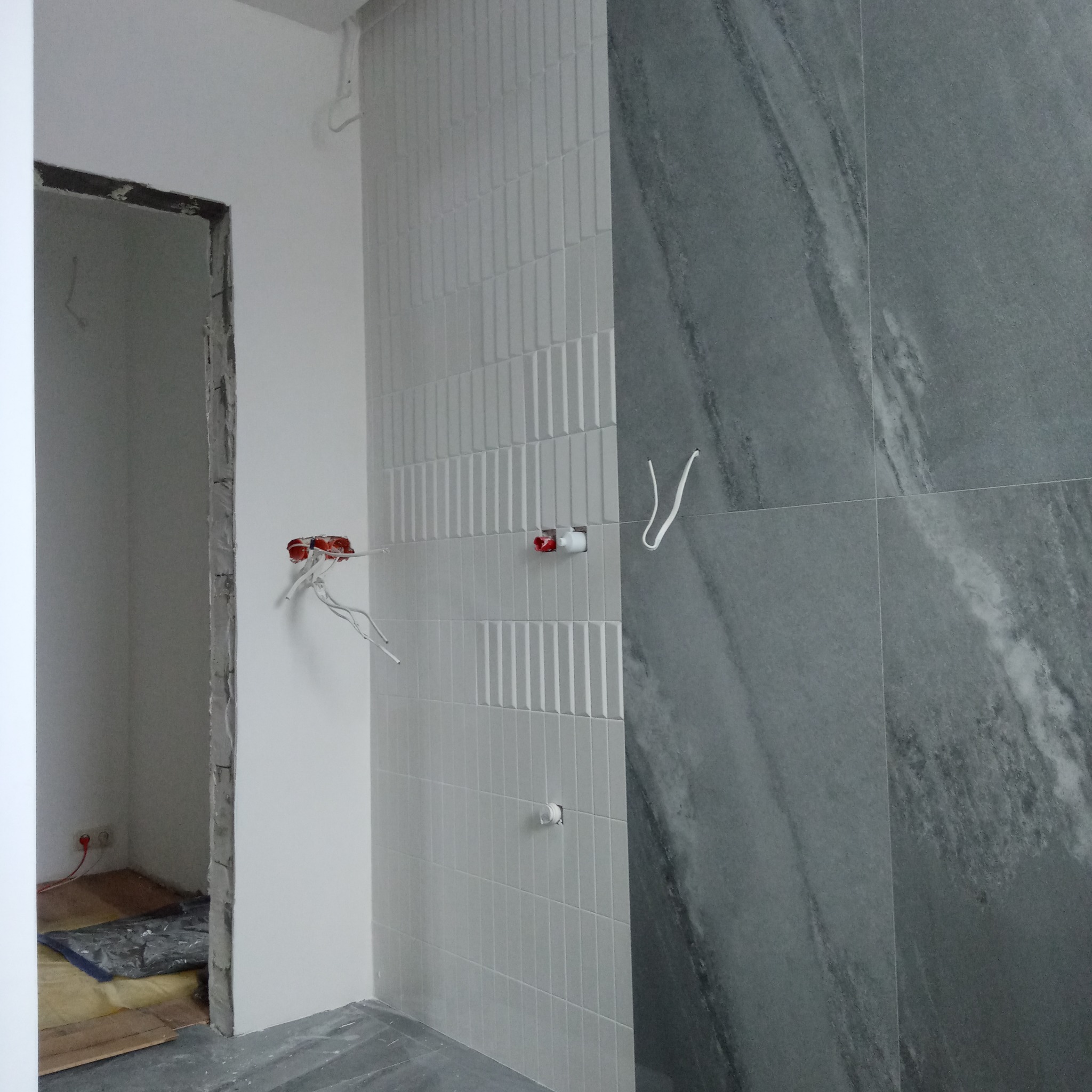 Wnętrze łazienki w trakcie remontu: białe, pionowe płytki ułożone w pasy obok szarych płyt imitujących marmur, wystające kable elektryczne, surowa ościeżnica drzwi.