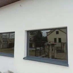 Dwa okna aluminiowe, prostokątne, grafitowe ramy, w białej, otynkowanej elewacji domu jednorodzinnego; w szybach odbija się sąsiedni budynek i drzewa.