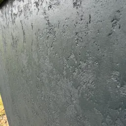 Szara ściana z wyraźną, nieregularną fakturą, widoczna z bliskiej perspektywy pod kątem, z fragmentem żółtej taśmy malarskiej w górnym rogu.