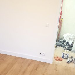 PAWLAN-PROJEKT Pavlo Kulbakov - Pokój po remoncie: Jasne panele podłogowe, białe ściany z listwami przypodłogowymi i gniazdkiem elektrycznym; w tle widoczne narzędzia i wiadro farby.