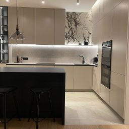 PAWLAN-PROJEKT Pavlo Kulbakov - Nowoczesna kuchnia w Warszawie z beżowymi szafkami, marmurowym akcentem na ścianie i czarnym barkiem z hokerami, oświetlona listwą LED podszafkową i wiszącą lampą ze szklanym kloszem.