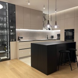 PAWLAN-PROJEKT Pavlo Kulbakov - Nowoczesna kuchnia z wyspą, beżowymi szafkami i czarnymi akcentami, oświetlona punktowymi lampami sufitowymi i wiszącymi lampami nad wyspą. Na blacie widoczne akcesoria kuchenne, a w przeszklonej...