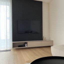 PAWLAN-PROJEKT Pavlo Kulbakov - Nowoczesny salon z czarną ścianą, telewizorem i beżową szafką RTV, uzupełniony o dekoracyjne, żłobkowane panele ścienne i drewnianą podłogę.
