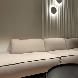 PAWLAN-PROJEKT Pavlo Kulbakov - Fragment salonu z beżową sofą modułową, okrągłym czarnym stolikiem kawowym i ścianą z pionowymi panelami oraz dwoma okrągłymi lampami LED.