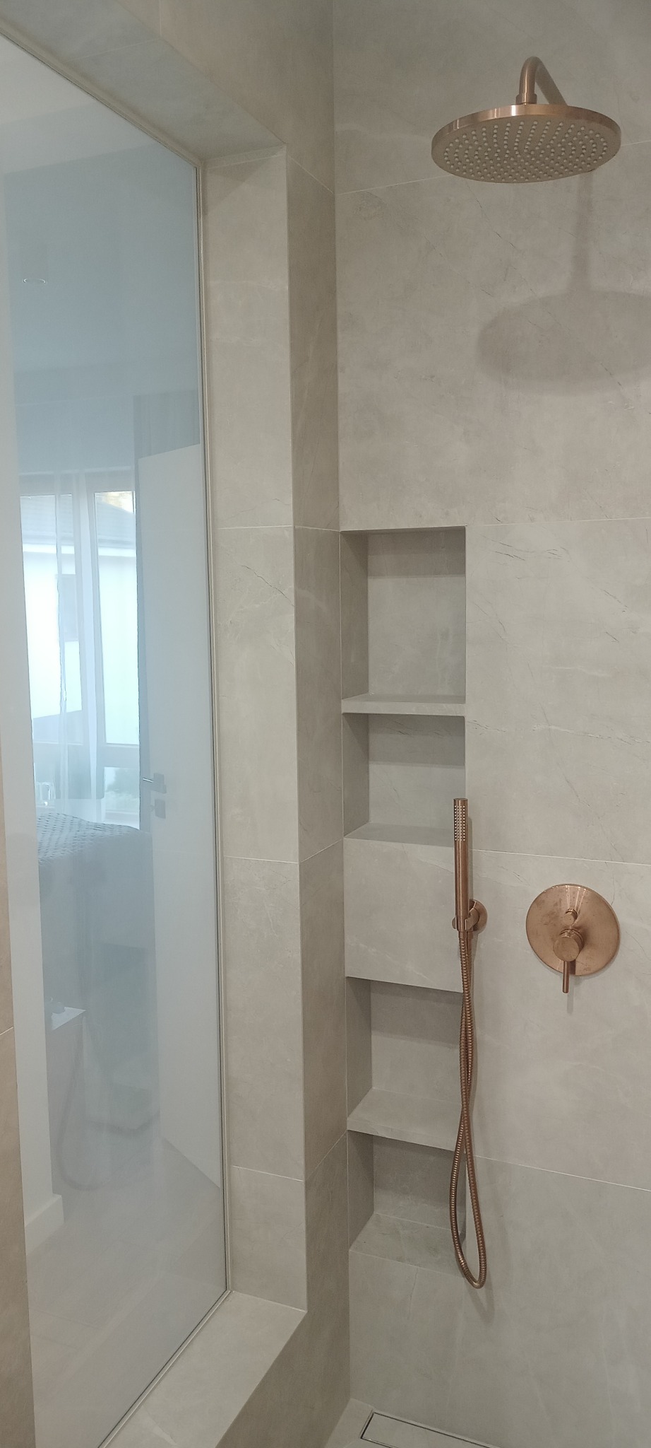 Nowoczesna łazienka z kabiną prysznicową, beżowe płytki imitujące marmur, wnęka z półkami, deszczownica i bateria w kolorze różowego złota.