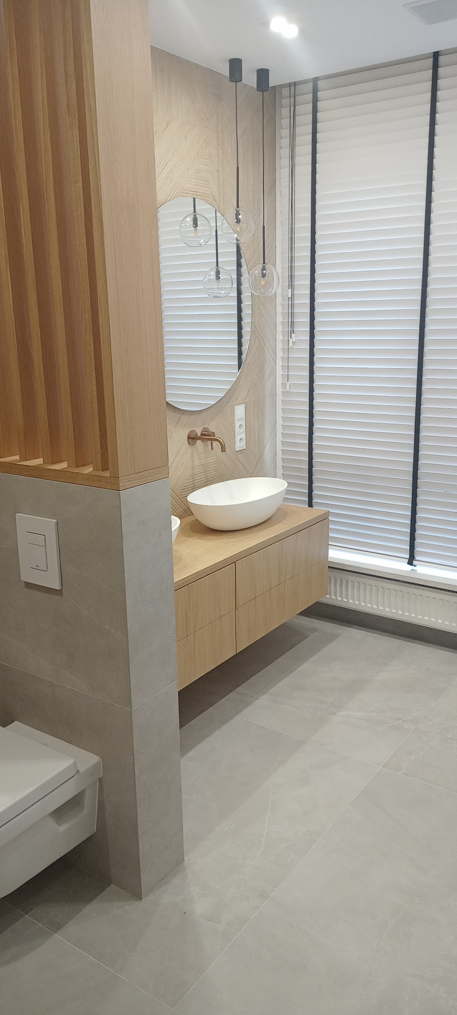 Nowoczesna łazienka z owalną umywalką nablatową, drewnianą szafką pod umywalką, okrągłym lustrem, armaturą w kolorze miedzi i trzema szklanymi lampami wiszącymi.