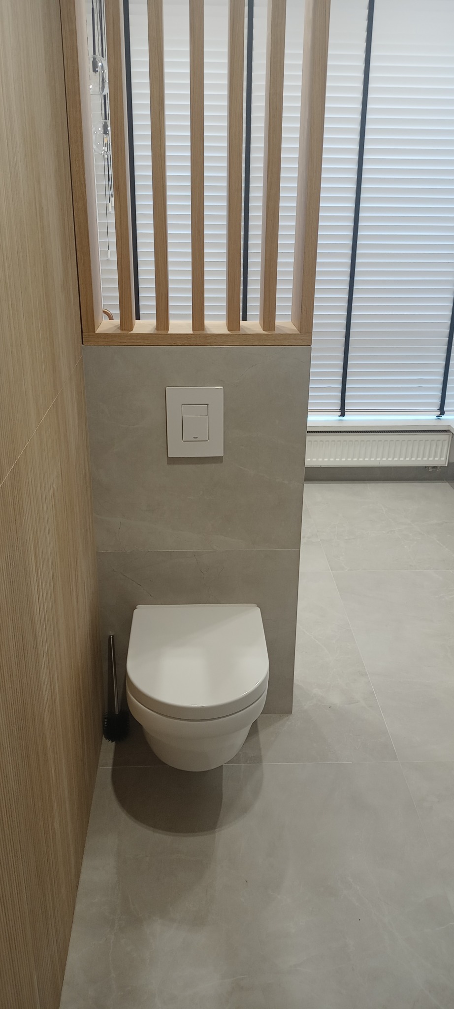 Nowoczesna toaleta wisząca w łazience wykończonej płytkami imitującymi marmur, z drewnianą ażurową ścianką działową i żaluzjami w oknie w tle.