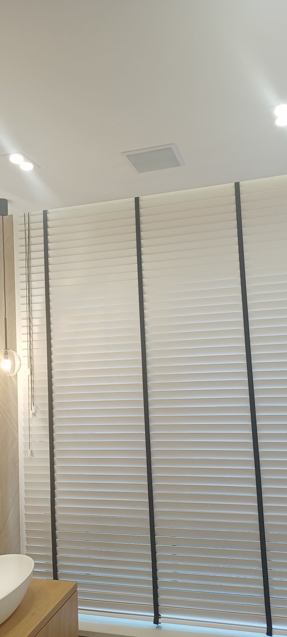 Zamknięte, białe żaluzje poziome z ciemnymi taśmami na tle białego sufitu z wbudowanym głośnikiem i oświetleniem punktowym, widoczny fragment umywalki i szafki łazienkowej.