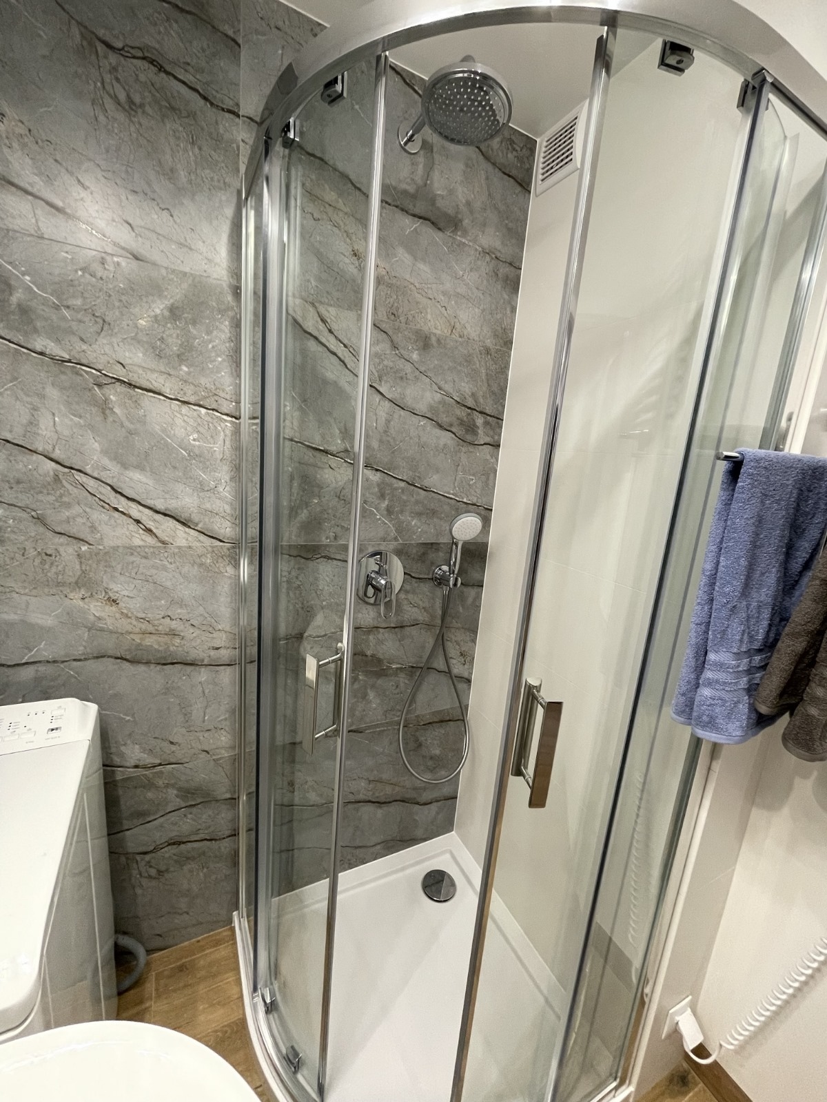 Narożna kabina prysznicowa z chromowanymi okuciami i deszczownicą, szare płytki imitujące marmur na ścianie, obok pralka i wiszące ręczniki.