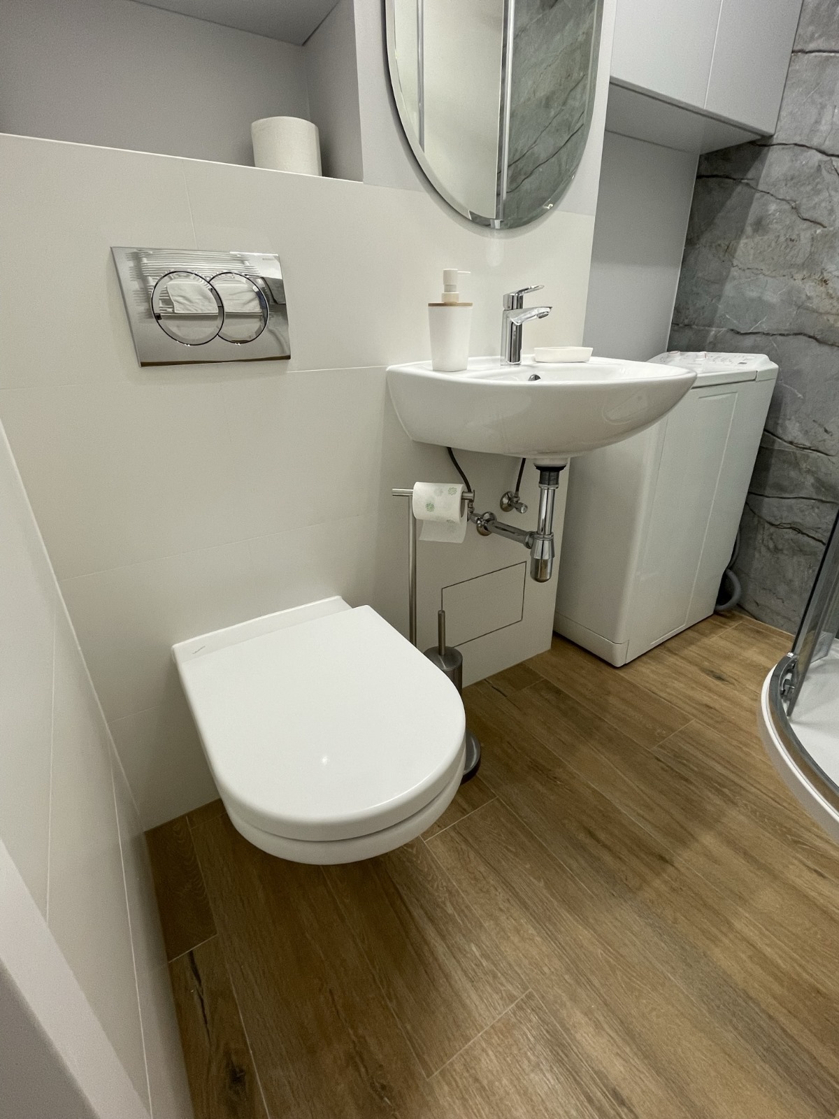 Nowoczesna łazienka z podwieszaną toaletą, umywalką nablatową, drewnopodobną podłogą i pralką w tle, oświetlona jasnym światłem.