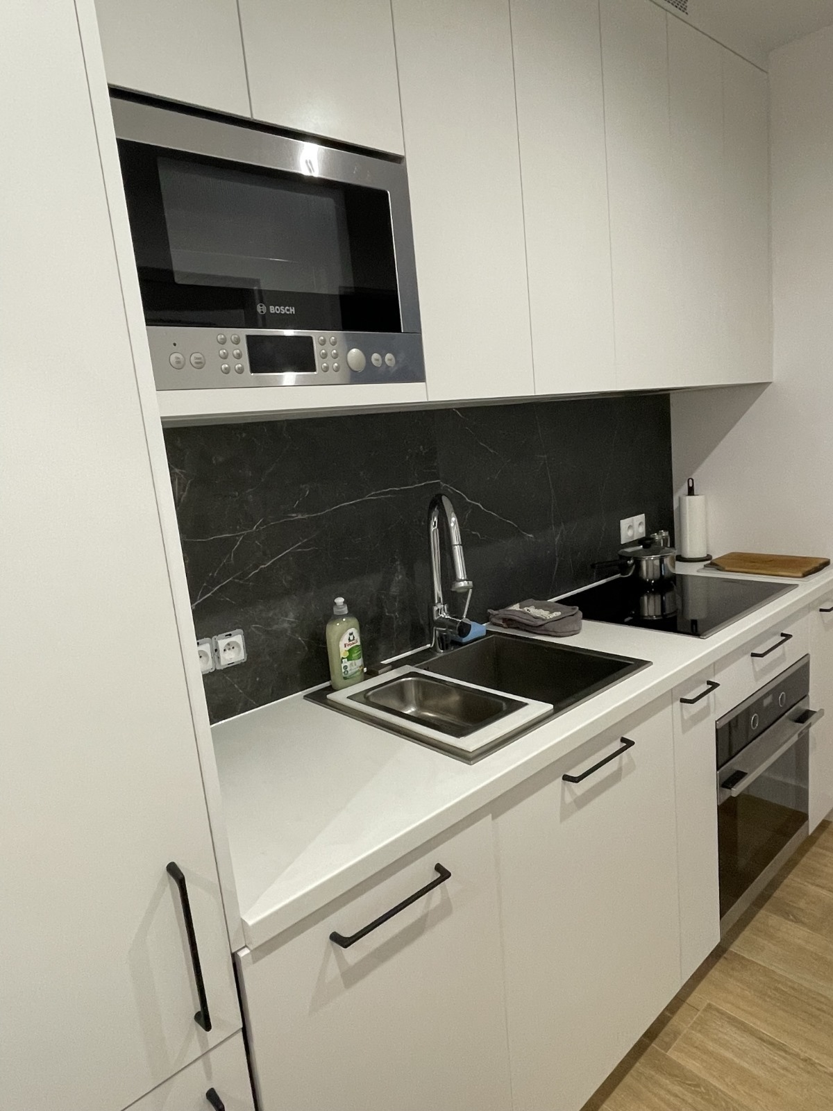 Nowoczesna, biała kuchnia z czarnym marmurkowym backsplashiem, wbudowanym mikrofalówką Bosch, czarnymi uchwytami szafek i płytą indukcyjną.