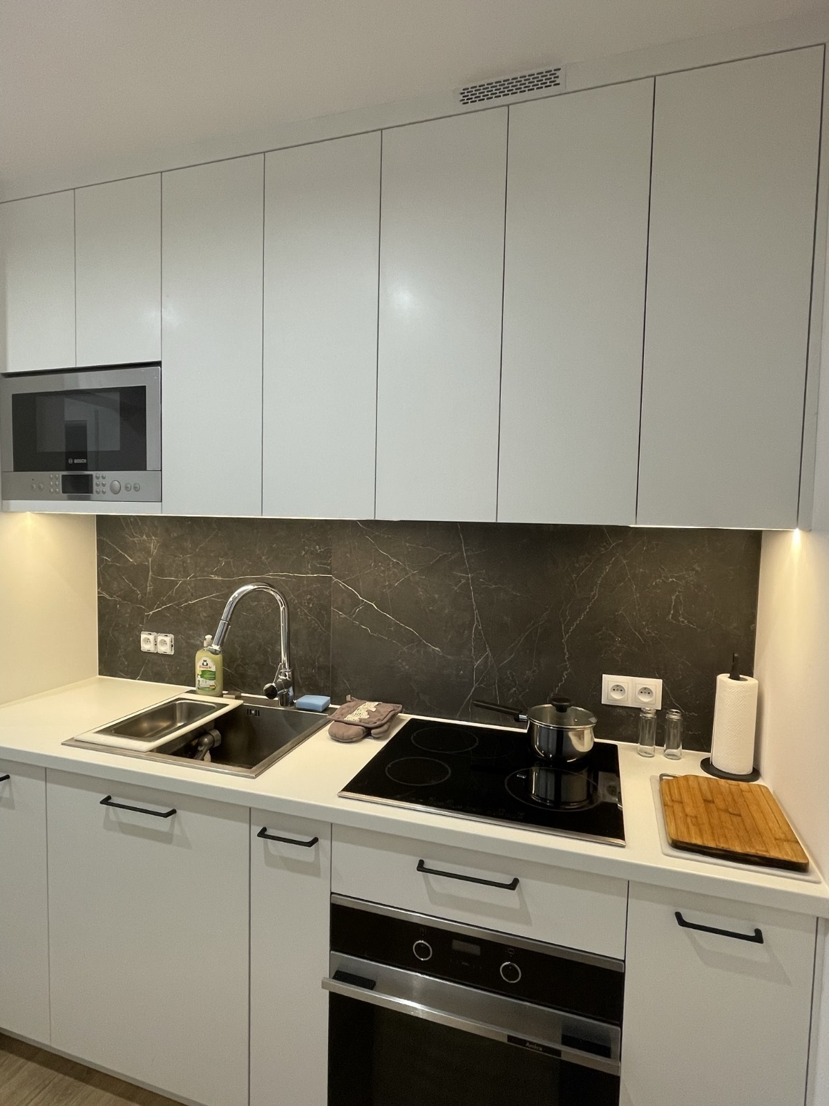 Nowoczesna kuchnia z białymi szafkami, czarnym marmurkowym backsplashiem, płytą indukcyjną z garnkiem i zlewem dwukomorowym.