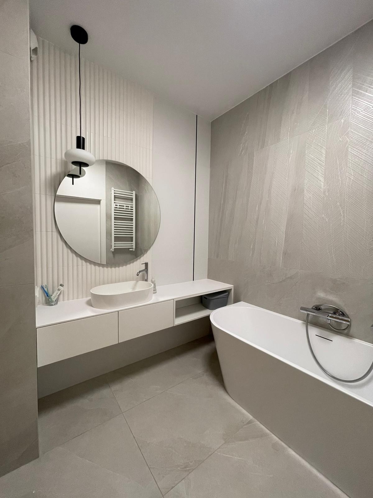 Nowoczesna łazienka z owalną umywalką nablatową, okrągłym lustrem, minimalistyczną lampą wiszącą i wolnostojącą wanną na tle szarych płytek z delikatnym wzorem.