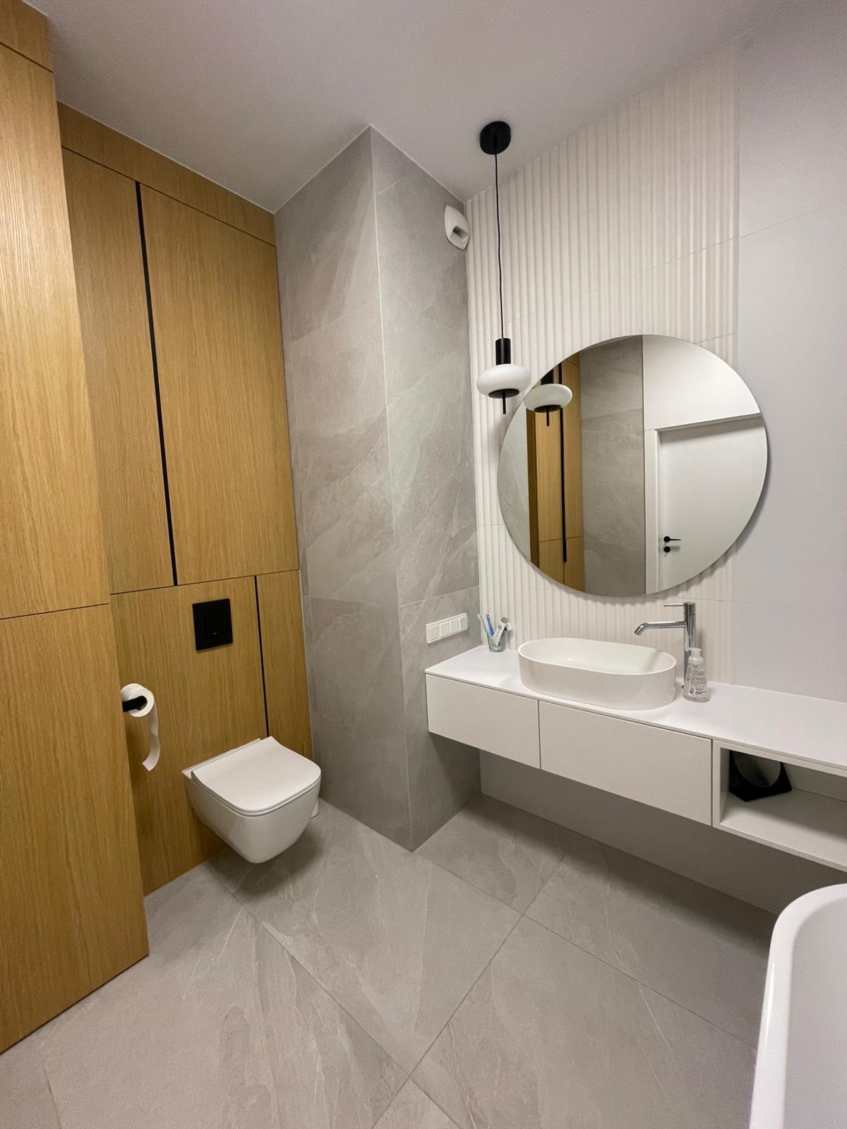 Nowoczesna łazienka z wiszącą toaletą, owalną umywalką nablatową, okrągłym lustrem i drewnianą zabudową, wykończona szarymi płytkami imitującymi kamień.