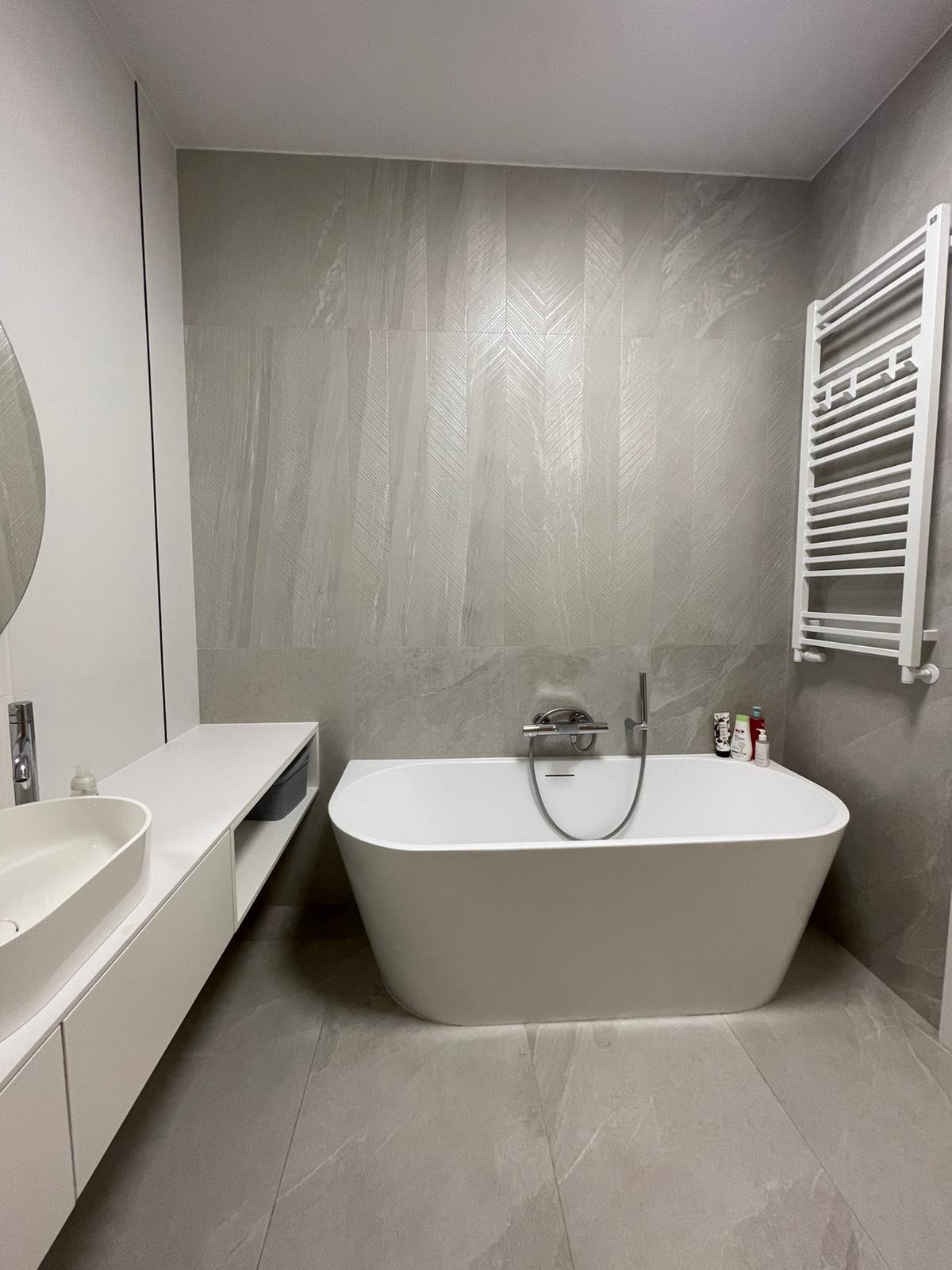 Nowoczesna łazienka w odcieniach szarości z wolnostojącą wanną, grzejnikiem drabinkowym i umywalką nablatową na długim blacie.