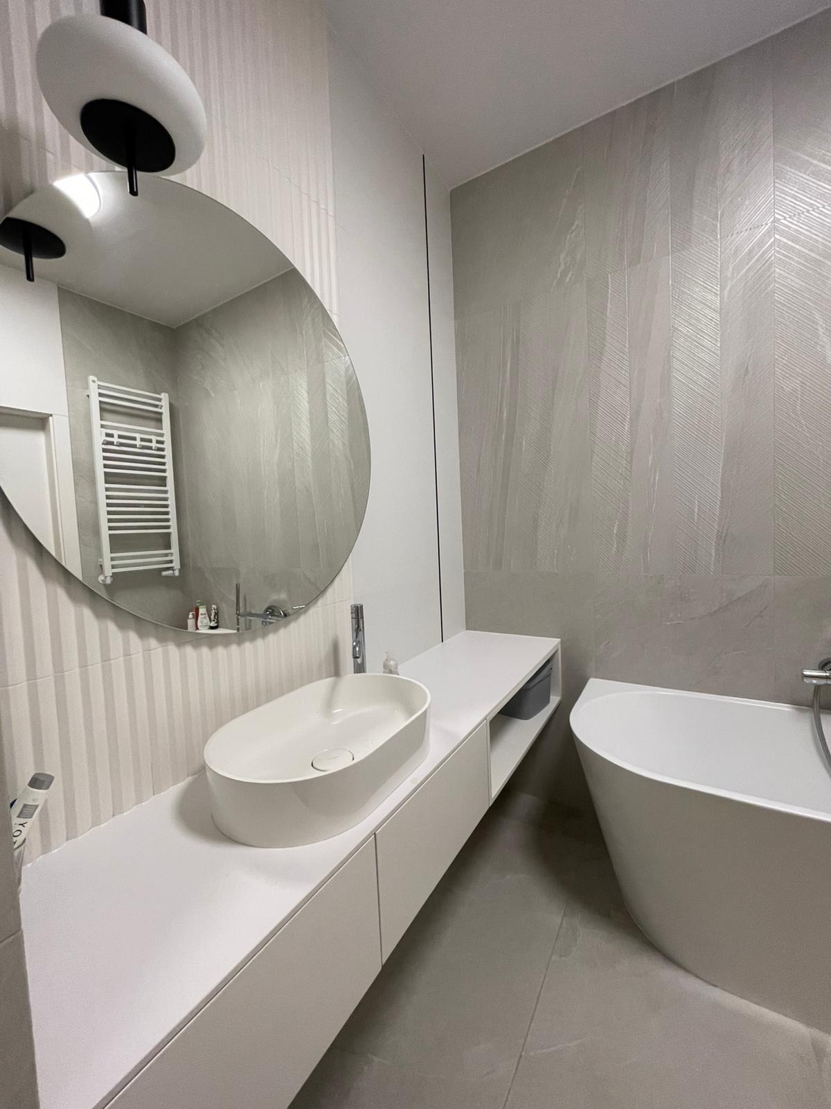 Nowoczesna łazienka w odcieniach szarości i bieli z owalną umywalką nablatową, asymetrycznym lustrem i wanną wolnostojącą, ściany wyłożone płytkami imitującymi naturalny kamień z subtelnym wzorem...