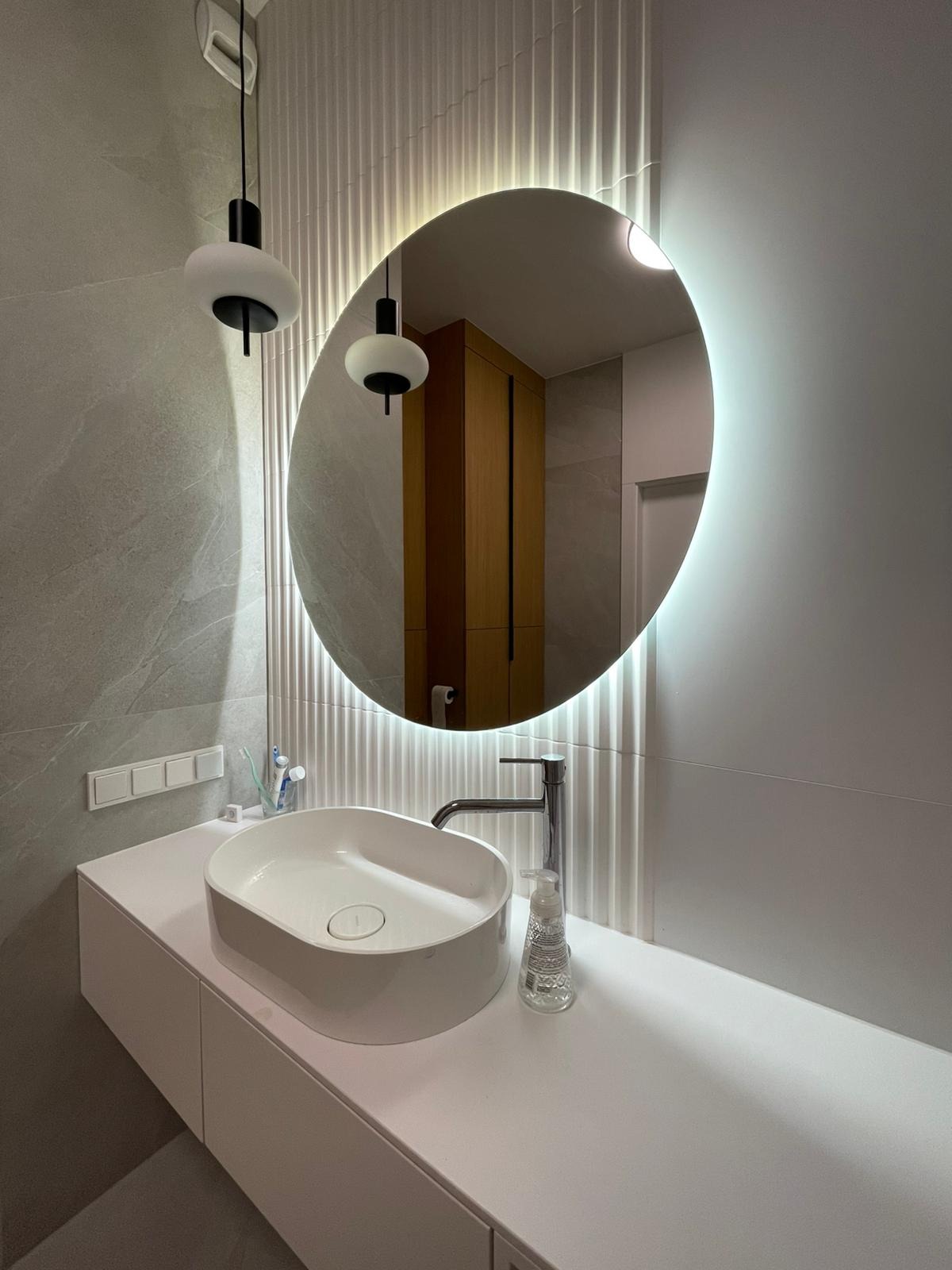 Nowoczesna łazienka z owalnym lustrem z podświetleniem LED, białą umywalką nablatową i baterią, szare płytki imitujące marmur na ścianie oraz wiszące lampy z białymi kloszami.