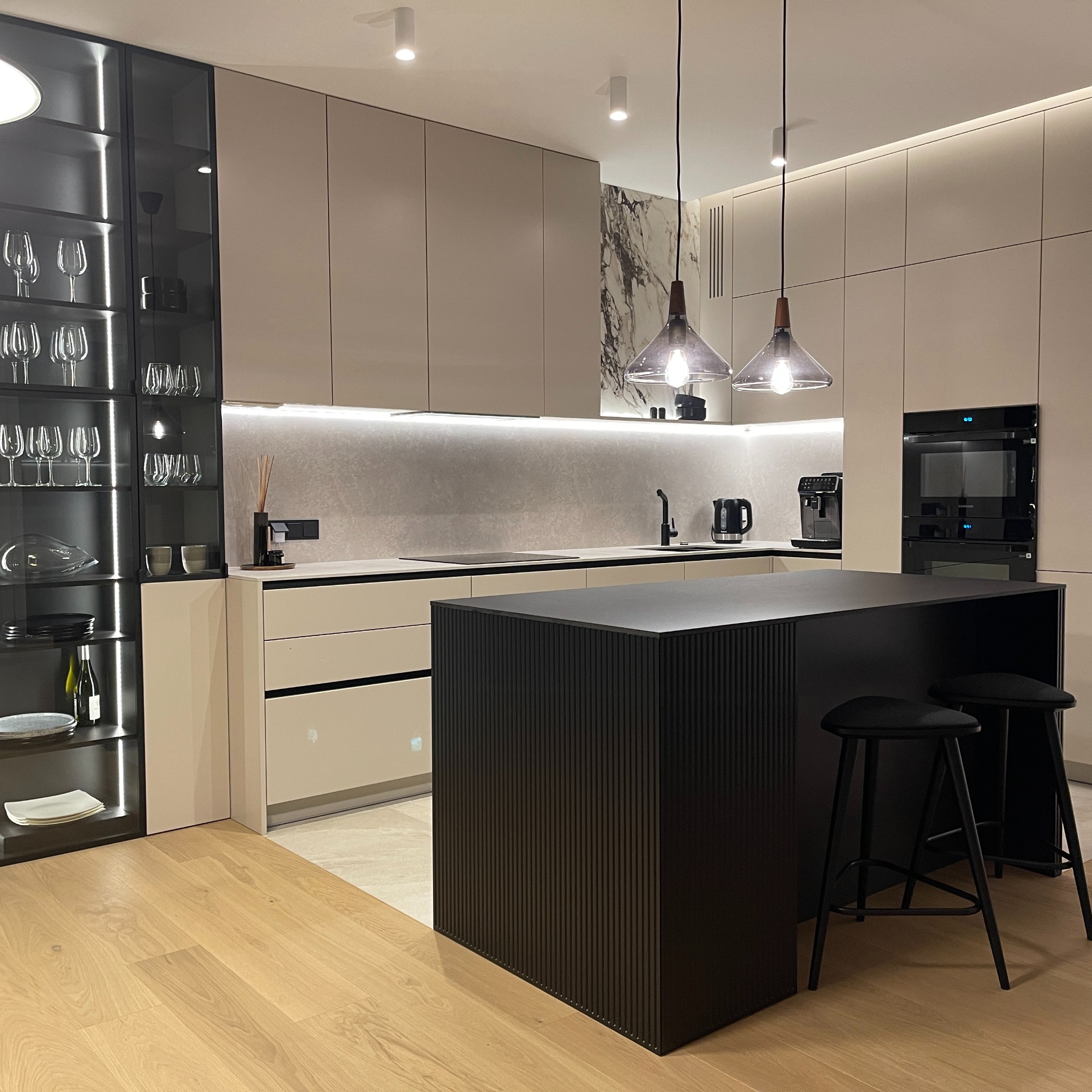 Nowoczesna kuchnia z wyspą, beżowymi szafkami i czarnymi akcentami, oświetlona punktowymi lampami sufitowymi i wiszącymi lampami nad wyspą. Na blacie widoczne akcesoria kuchenne, a w przeszklonej...